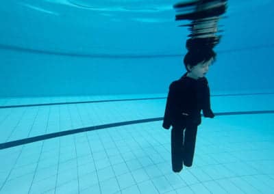 Child in dark clothing underwater in a pool.