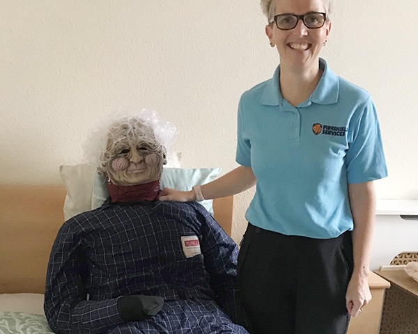 Caregiver with elderly resident dummy in care facility room.