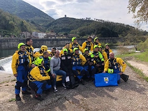 Rescue team gathers beside river with mountainous backdrop.